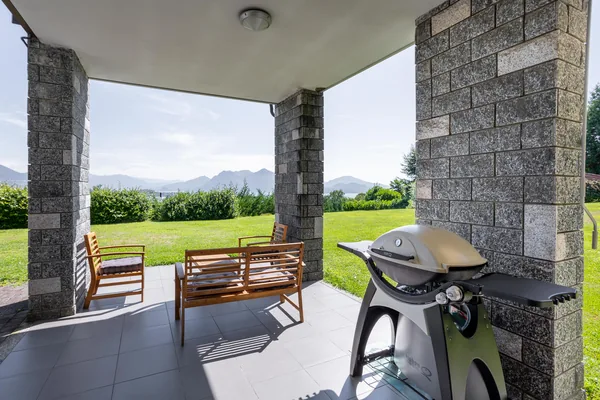 Covered stone portico with armchairs and a gas barbecue, looking over the lawn to the lake